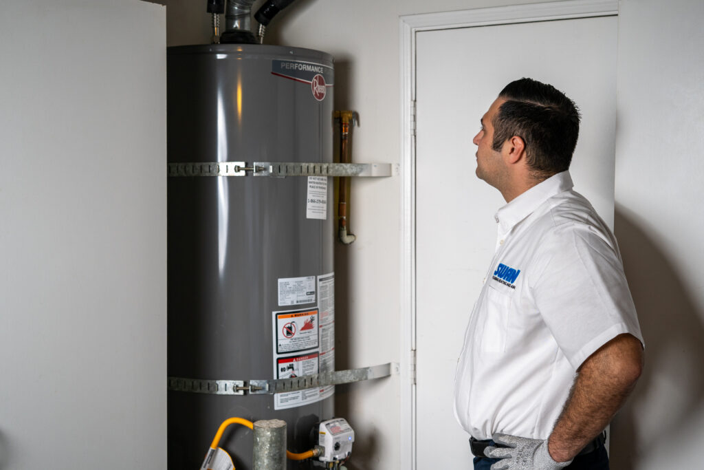 A Swan Plumbing, Heating, and Air technician in uniform inspects a gray water heater in a clean, organized utility room.
