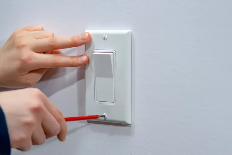 A Swan Plumbing, Heating, and Air technician carefully unscrews the bottom of a white light switch plate with a red screwdriver.