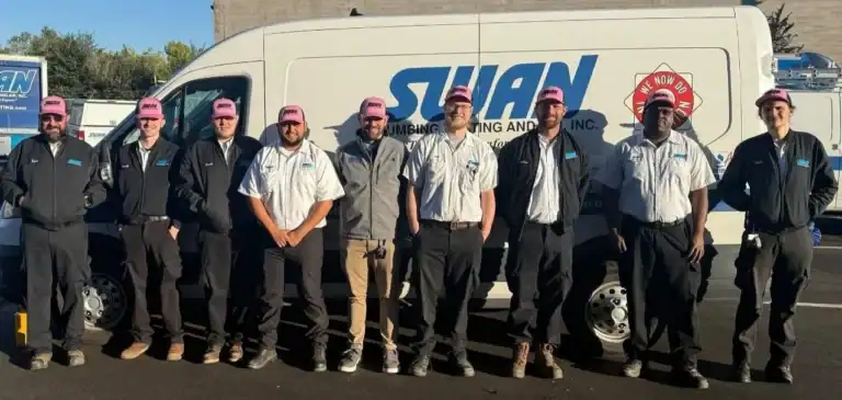 A team of nine Swan Plumbing, Heating and Air professionals, dressed in work uniforms and pink hats, stand together in a sunny parking lot in front of their company van, ready to provide reliable service.