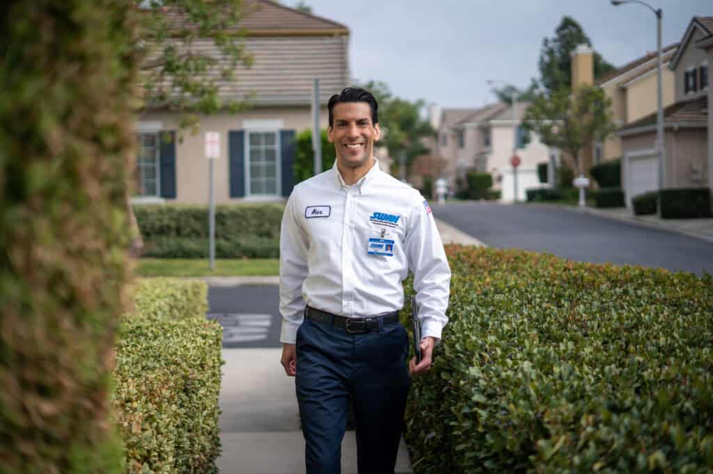 A friendly Swan Plumbing, Heating, and Air technician, dressed in uniform, walks along a Denver, CO residential sidewalk after completing a professional thermostat replacement for a customer.