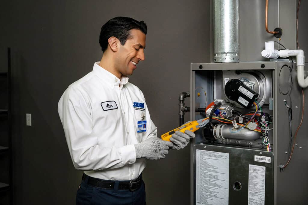 A Swan Plumbing, Heating, and Air technician in uniform uses a digital multimeter to carefully test an HVAC system, with the furnace panel open in a tidy utility room.
