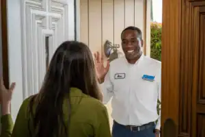 A friendly Swan Plumbing, Heating, and Air technician in a crisp white uniform and name tag stands at the front door, warmly waving to a customer with long hair who has just answered. The technician is ready to help resolve a breaker panel tripping issue during the fall season in Denver, CO.